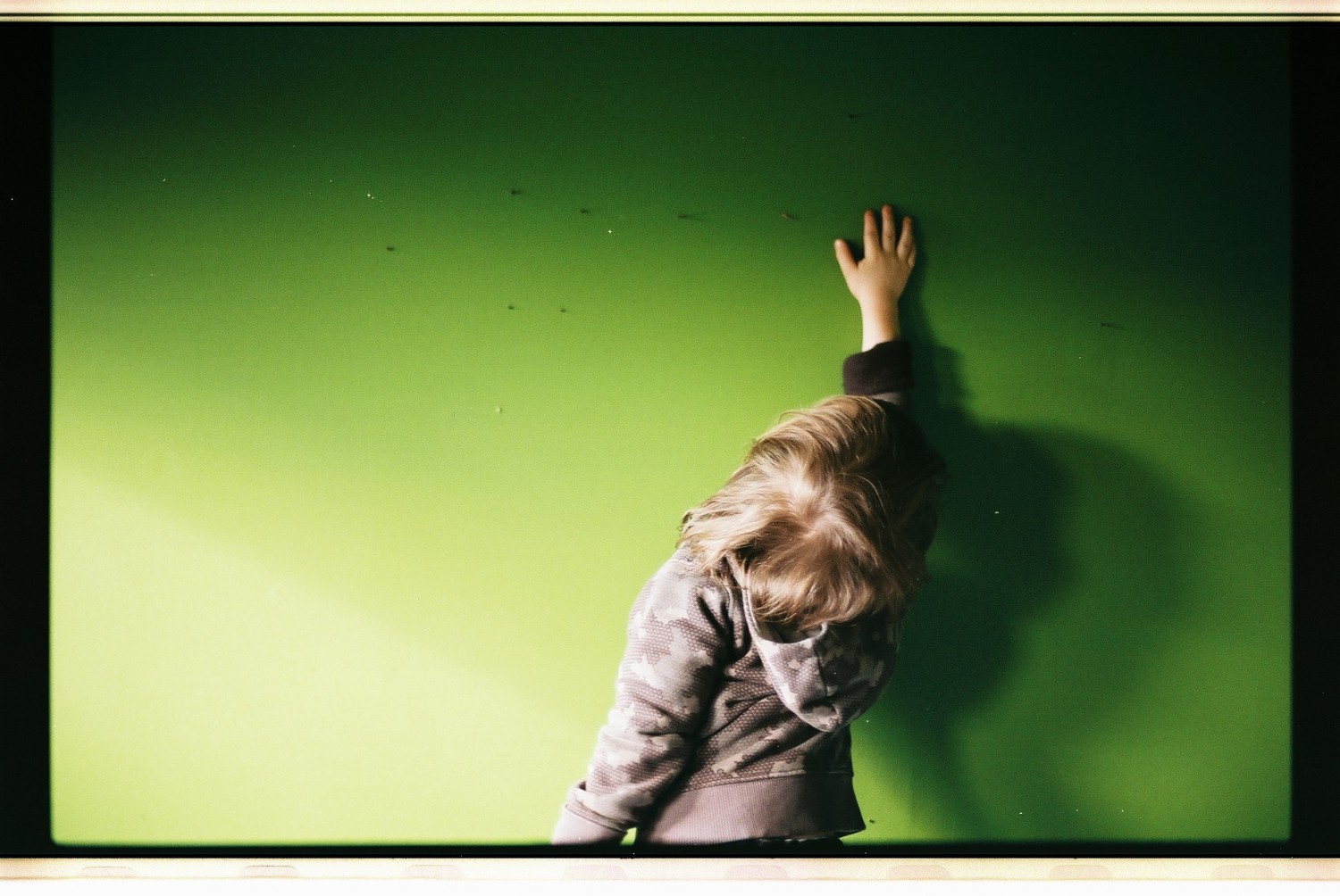 Dziecko z wyciągniętą do góry ręką na tle zielonej ściany.